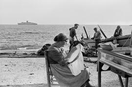 Concepción Jiménez Heredia (Concha la de la lejía) bordando en la playa de La Malagueta. 6 de octubre de 1954. Málaga, España. A la derecha, la barca Trinidad. Al fondo, el buque Caronia